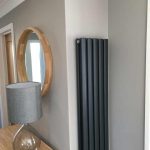 Wall with vertical radiator, round mirror and side table with table lamp.